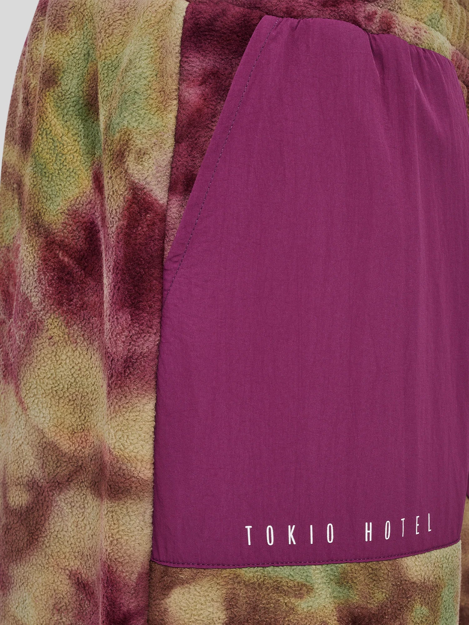Nahaufnahme der Hose mit kontrastierendem, violettem Nylon-Einsatz auf Batikfleece in Grün, Braun und Bordeaux. Auf dem Einsatz ist der weiße „TOKIO HOTEL“-Schriftzug aufgedruckt.