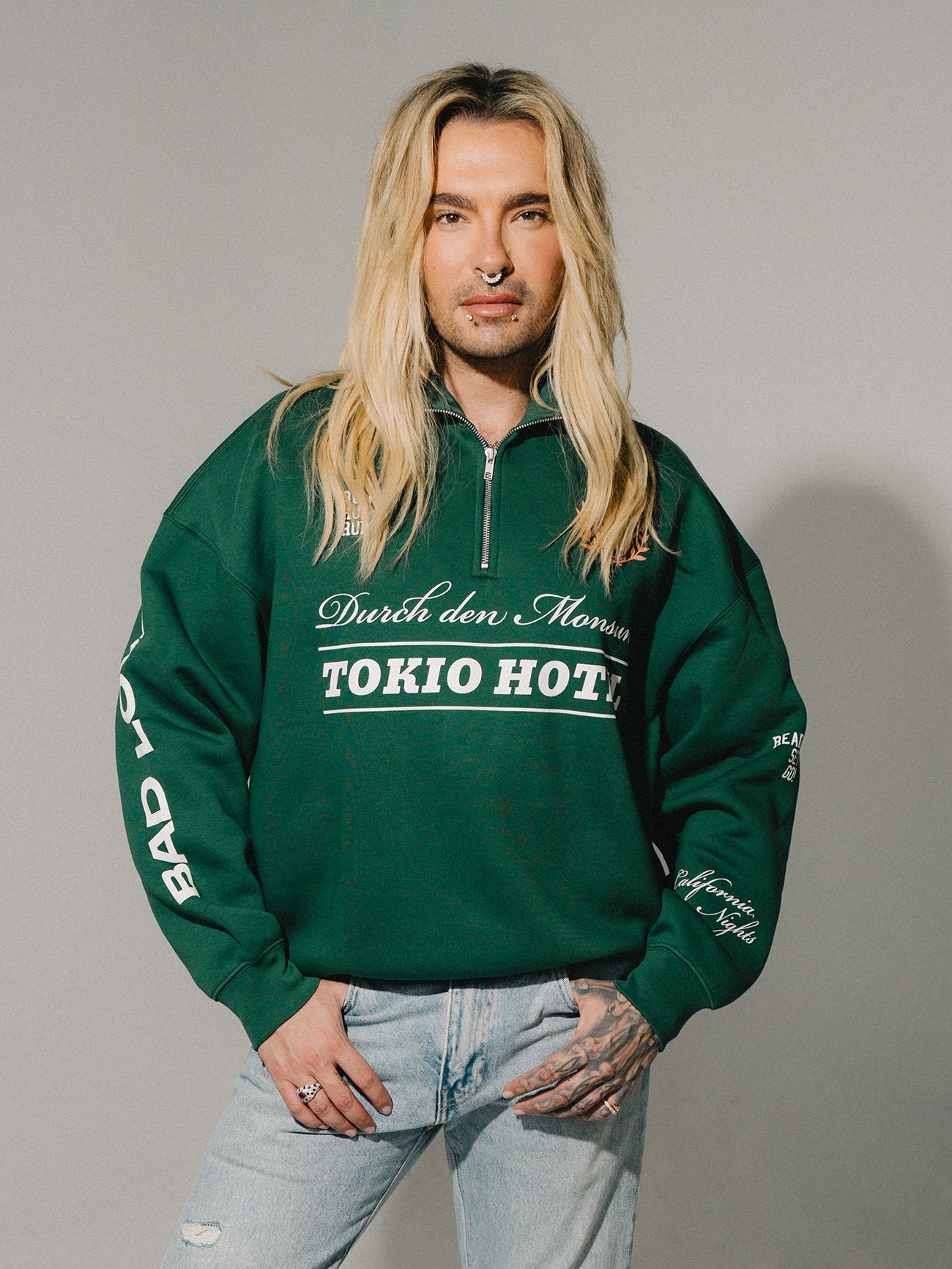 Person mit langen blonden Haaren trägt einen grünen Half-Zip-Sweater mit „Durch den Monsun“ und „TOKIO HOTEL“-Print, kombiniert mit heller Jeans.