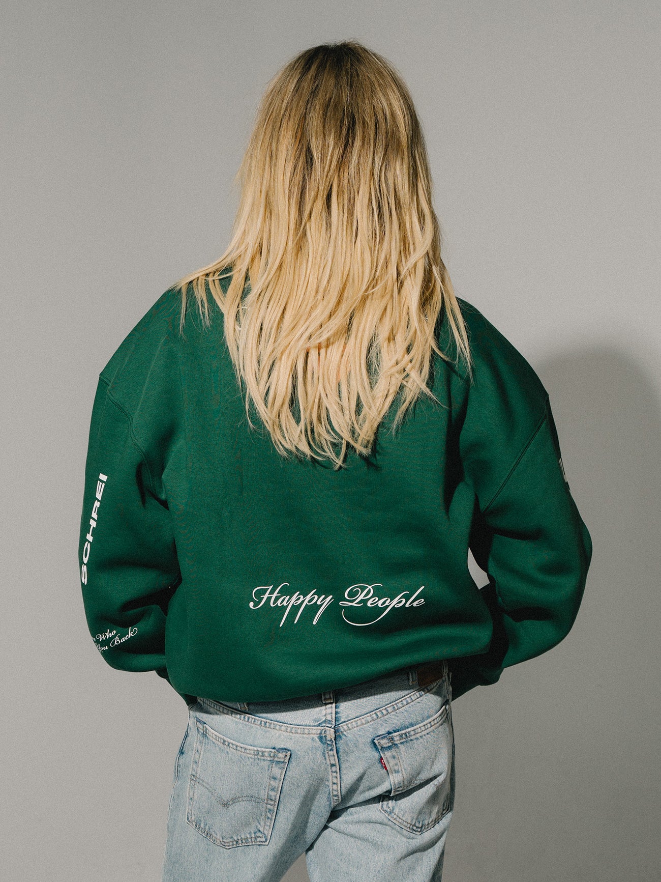Rückansicht einer Person im grünen Sweater mit „Happy People“-Schriftzug auf dem Rücken und heller Jeans.