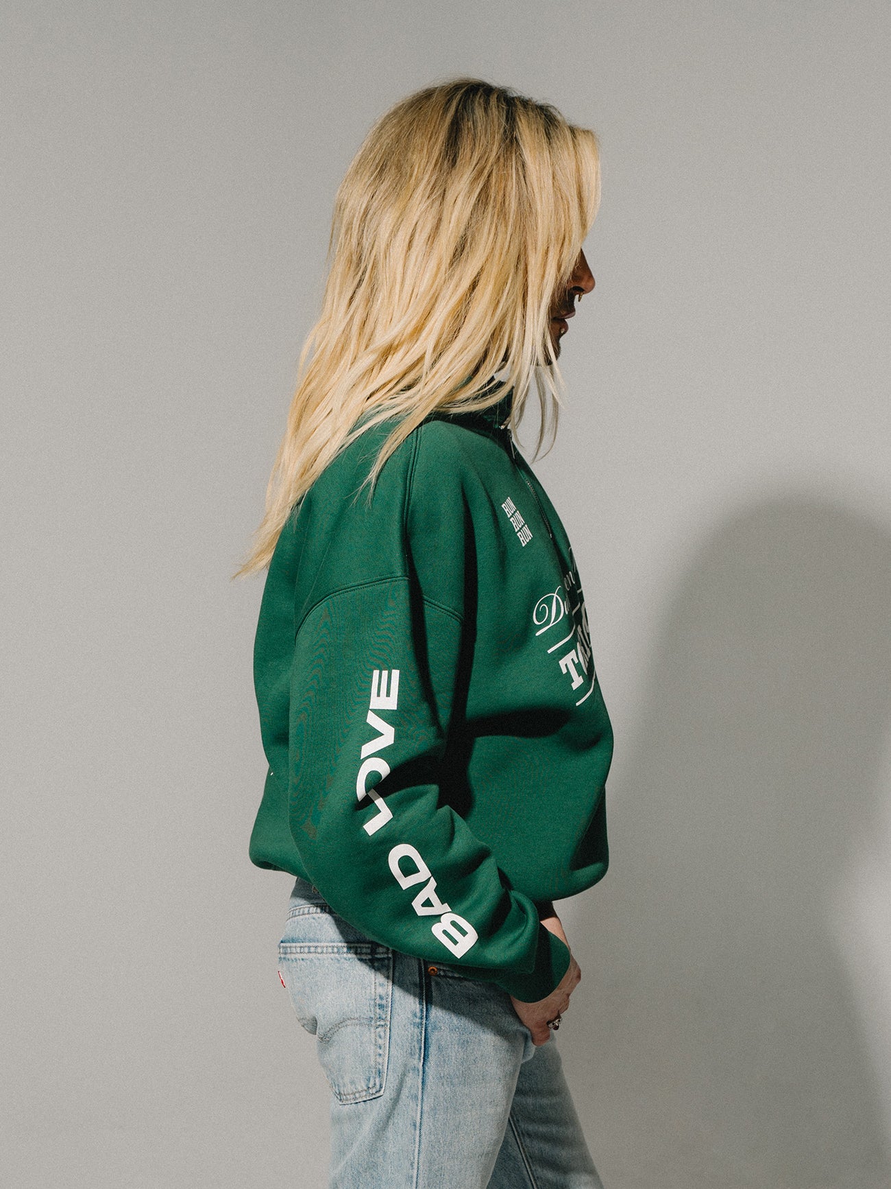 Seitenansicht einer Person im grünen Sweater mit „BAD LOVE“-Print auf dem Ärmel und hellem Denim.