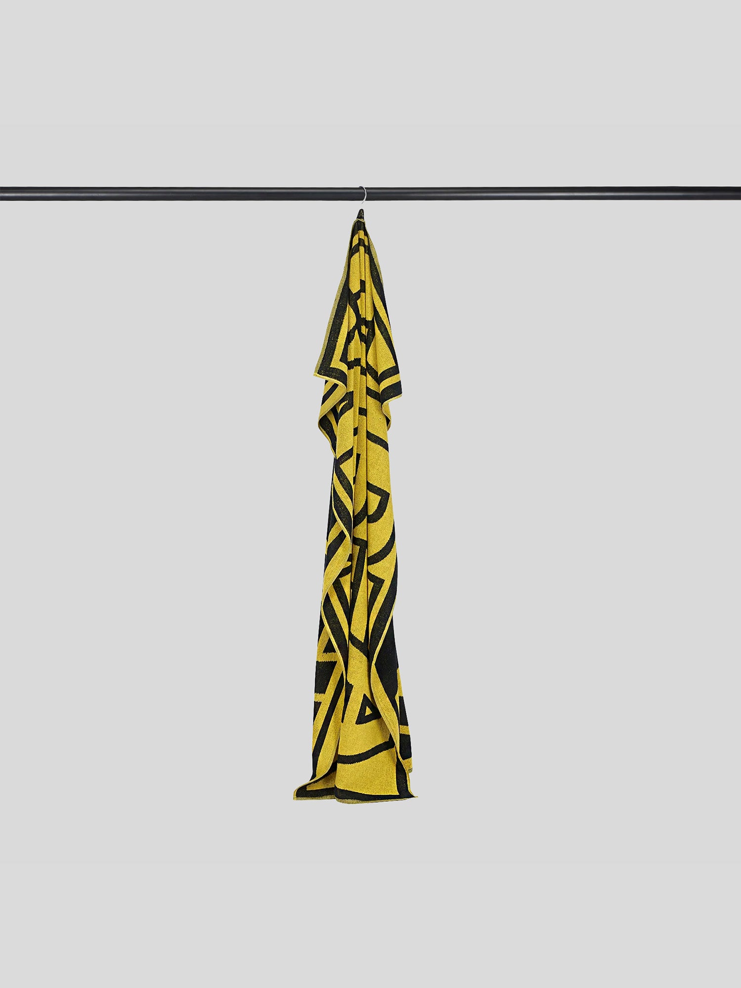 Das gelb-schwarze MDLA-Handtuch hängt locker gefaltet an einem schwarzen Metallhaken. Die kontrastreiche Typografie ist nur angedeutet sichtbar.