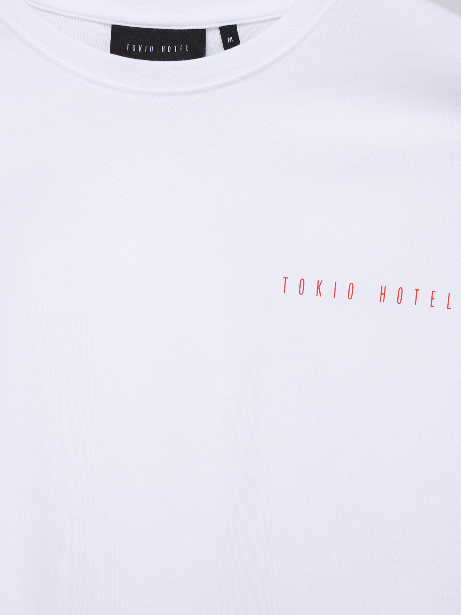 Nahaufnahme des oberen Brustbereichs eines weißen T-Shirts mit schmalem, roten „TOKIO HOTEL“-Schriftzug in Großbuchstaben. Das Innenlabel im Nacken ist ebenfalls sichtbar.