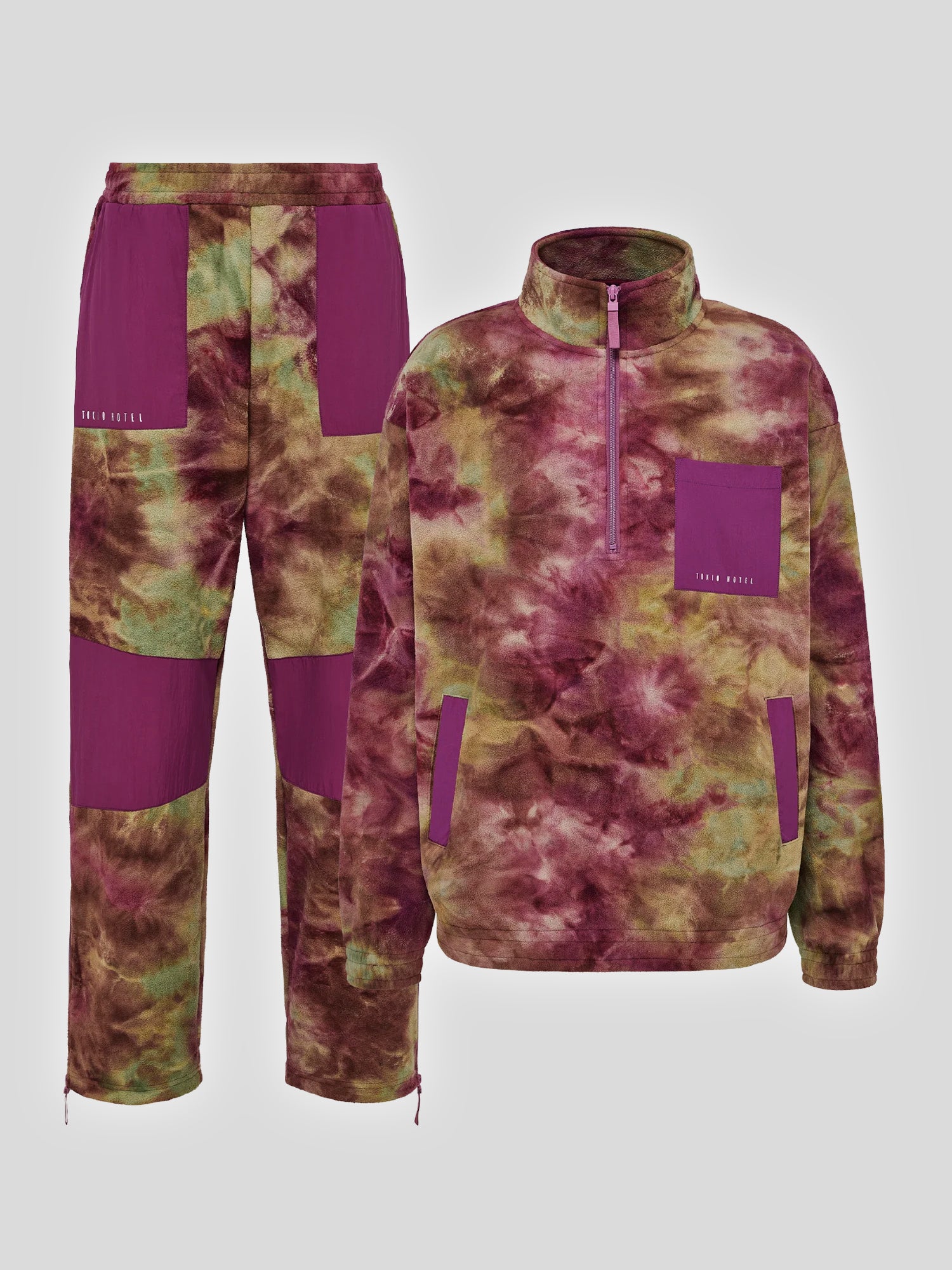 Zweiteiliges Freizeit-Outfit bestehend aus einer Hose und einem halben Zip-Fleece in Batik-Optik. Das Material zeigt ein Muster in Bordeauxrot, Olivgrün und Braun. Die Hose hat auffällige violette Kontrasteinsätze an den Oberschenkeln und Knien sowie einen elastischen Bund. Die Jacke besitzt einen kurzen Reißverschluss, eine violette Brusttasche und seitliche Eingrifftaschen. Beide Kleidungsstücke sind mit einem kleinen „TOKIO HOTEL“-Schriftzug versehen.