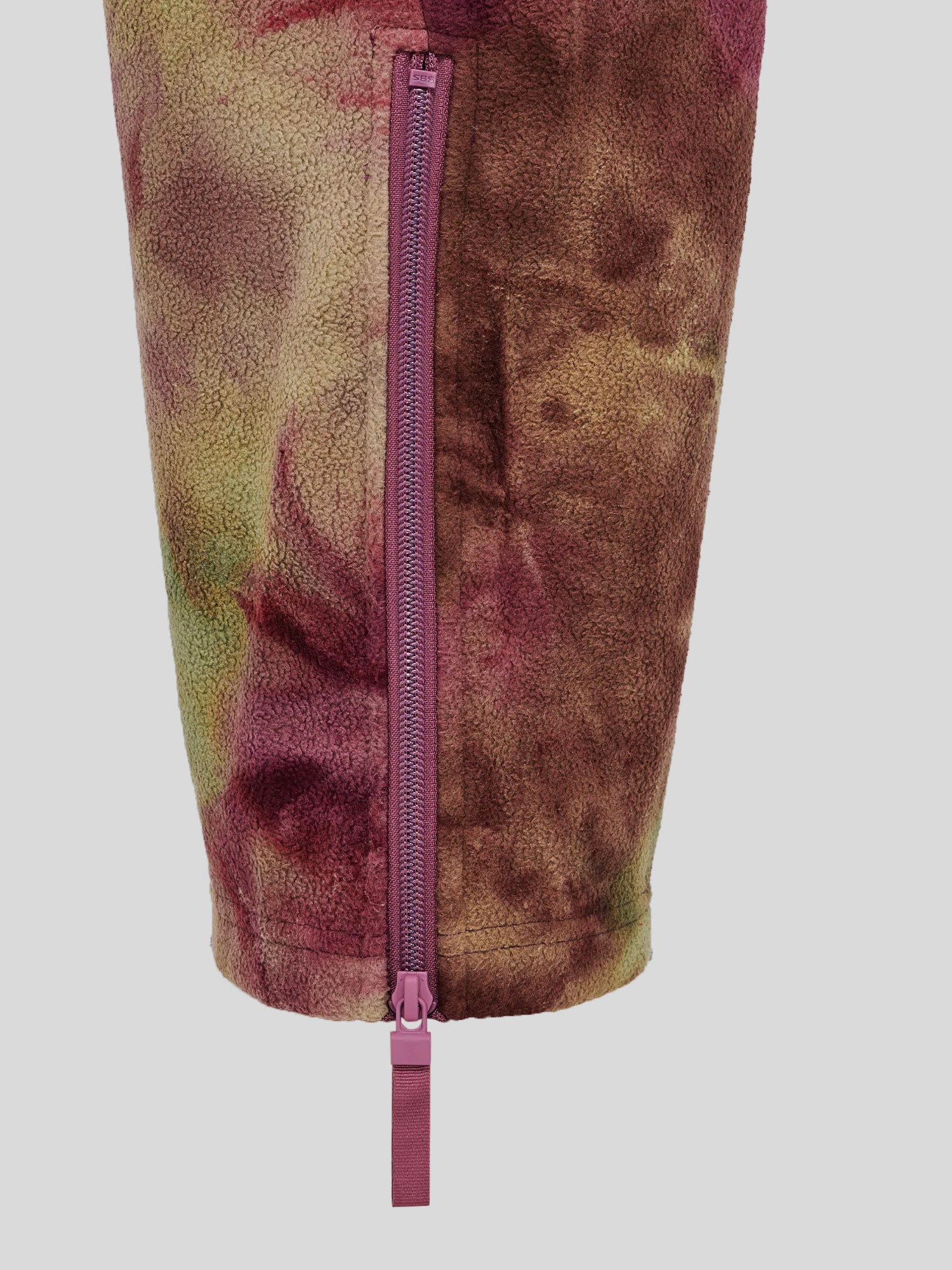 Nahaufnahme des Bein- bzw. Ärmelabschlusses mit seitlichem Reißverschluss in Violett. Der Zipper ist mit einem breiten Zugband versehen, der Stoff zeigt ein mehrfarbiges Batikmuster.