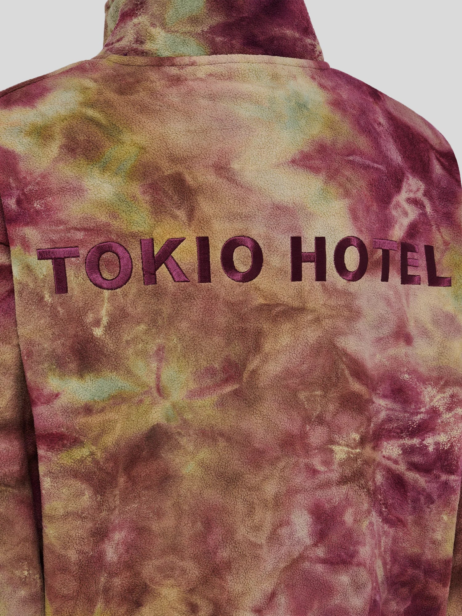 Detailaufnahme der Rückseite eines Batik-Fleecepullovers mit großflächigem, aufgesticktem „TOKIO HOTEL“-Schriftzug in dunklem Violett auf mehrfarbigem Grund in Bordeaux, Olive und Braun.
