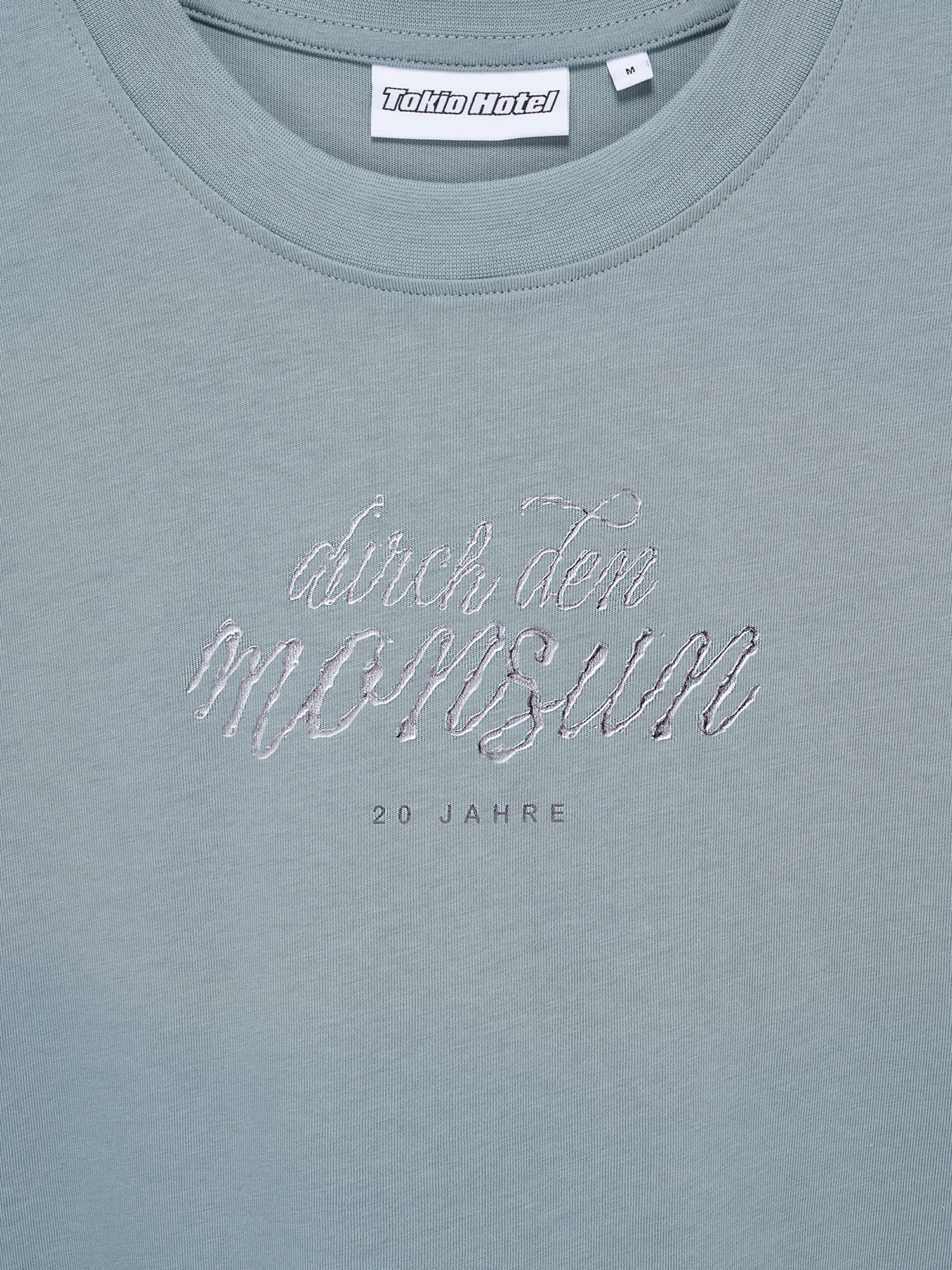 Nahaufnahme des gestickten Schriftzugs „durch den Monsun“ und der darunter gesetzten Worte „20 Jahre“ auf hellblauem Stoff.