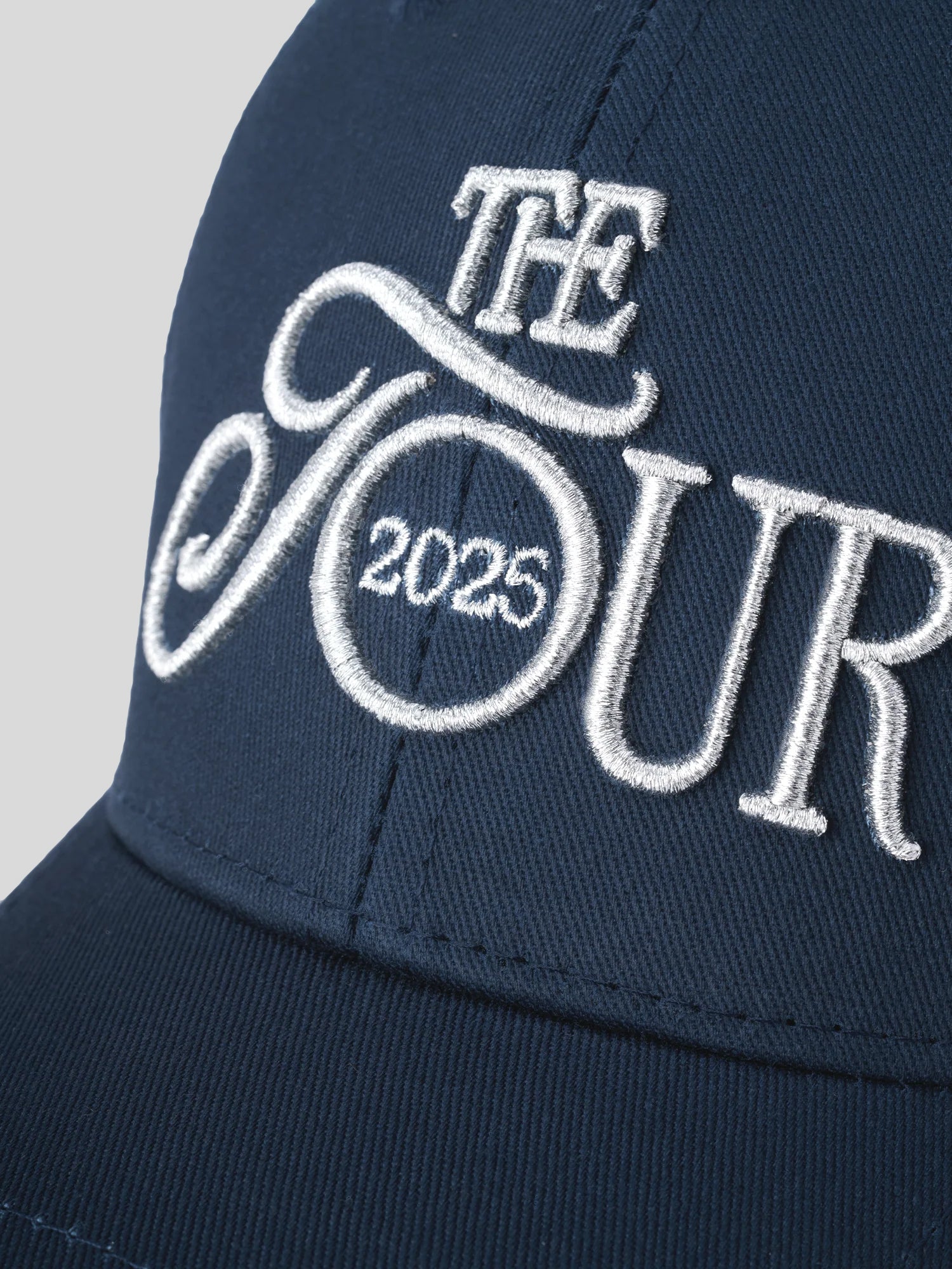 Dunkelblaue Cap mit aufgesticktem weißem Schriftzug „THE TOUR 2025“ in geschwungener Typografie, Nahaufnahme der detaillierten Stickerei auf der Vorderseite.