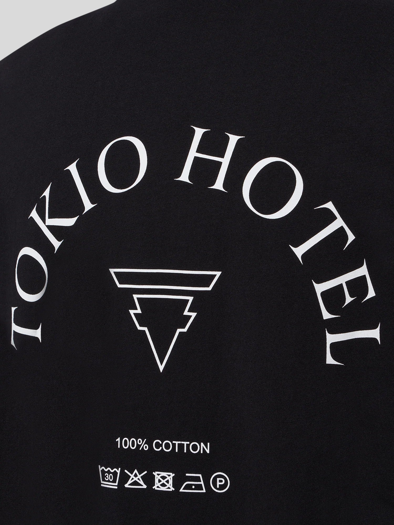 Nahaufnahme des schwarzen Longsleeves mit weißem Halbkreis-Schriftzug „TOKIO HOTEL“, stilisiertem Band-Logo darunter sowie Pflegehinweisen und „100% COTTON“-Angabe.