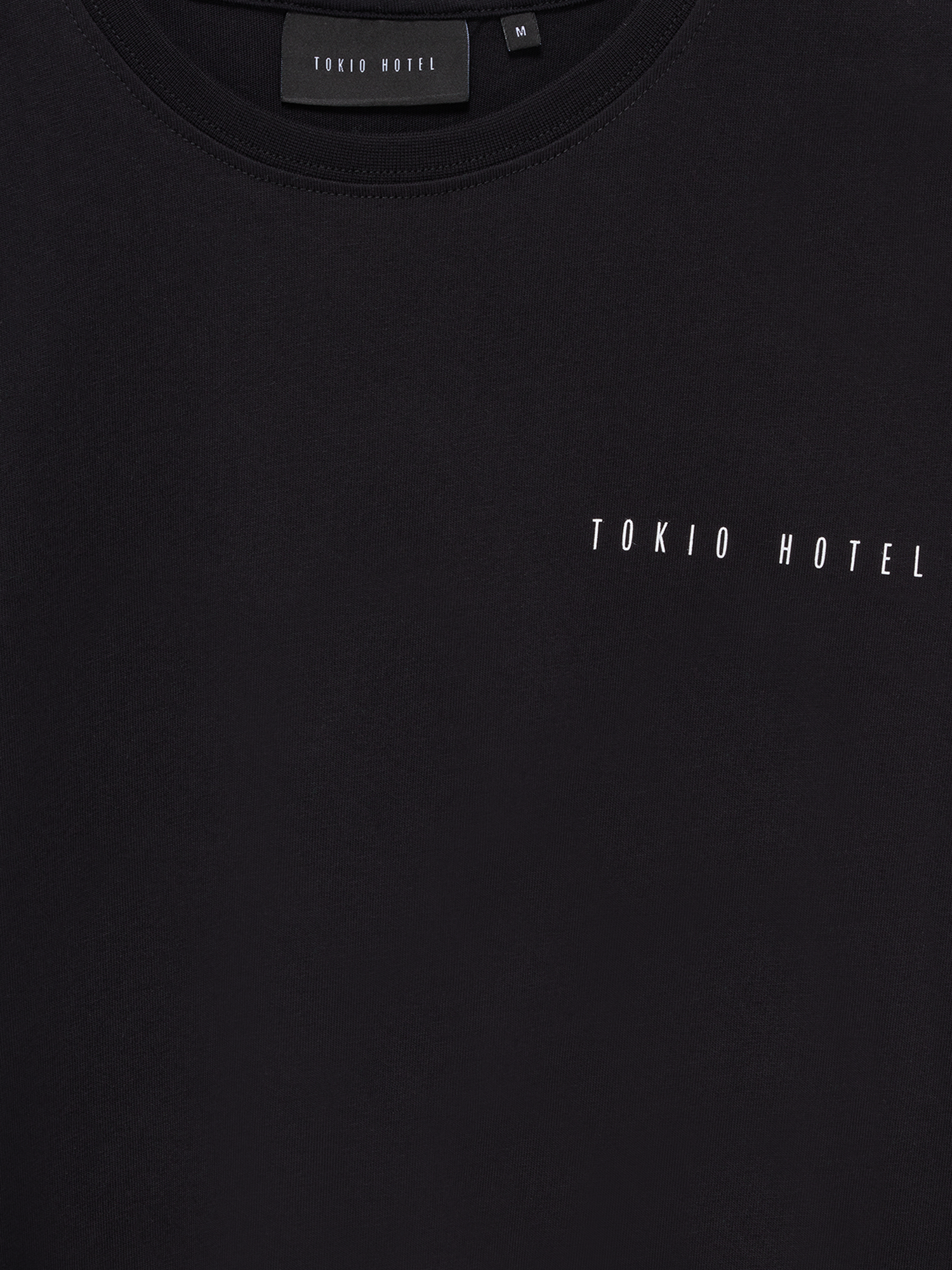 Nahaufnahme des oberen Brustbereichs eines schwarzen T-Shirts mit feinem, weißen „TOKIO HOTEL“-Schriftzug in schmaler Großbuchstabenschrift. Sichtbares Innenlabel im Nacken.